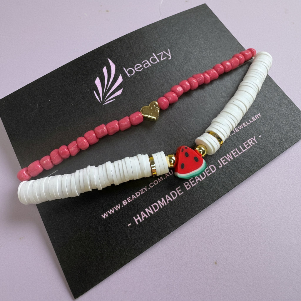 Two beaded bracelets, one red and one white with a watermelon charm, on a black card with branding on a yellow background. - Beadzy
