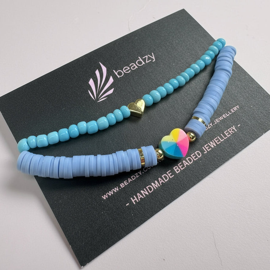 Blue beaded bracelet with a colorful heart charm on a black card with branding on a yellow background -Beadzy