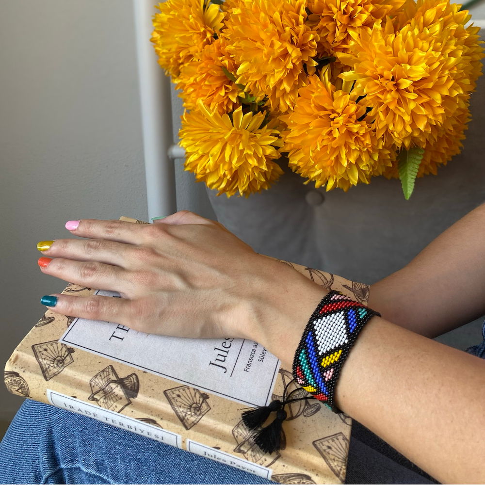 Person holding a bouquet of yellow flowers with a book underneath, wearing a colorful beaded bracelet.